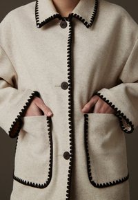 Beige villane coat musta äärisega, suured nuppude kinnitused ja kaks küljetaskut, nähtavad õmblusdetailid äärtes.