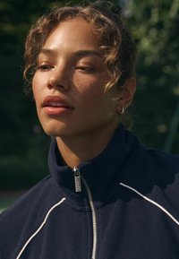 Tumesinine fliisist pullover kõrge kraega, lihvitud hõbedase luku ja valgete triipudega rinna kohal. Modellil on lokkis juuksed ja peened frecklid.