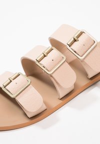 Sandali in pelle beige con tre fasce larghe, ciascuna con una fibbia dorata metallica, e una superficie superiore texturizzata. Suola piatta color tabacco.