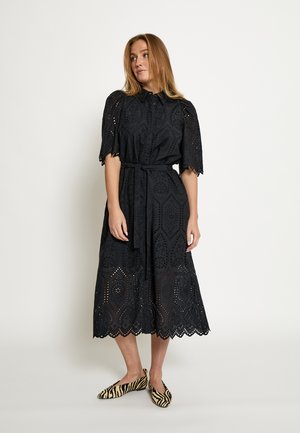 Zwarte midi-jurk van kant met korte mouwen, ingewikkelde uitsnijdingspatronen, een ceintuur met strik in de taille en een gerimpelde zoom. Gecombineerd met gestreepte platte schoenen.