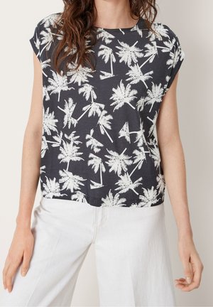 Frau trägt ein schwarzes Kurzarmshirt mit weißem Palmenmuster und hoch taillierte weiße Hose, steht vor einem neutralen Hintergrund.