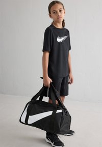 Svart treningst-skjorte og shorts med hvit Nike-logo, holder en svart duffelbag med hvit Nike-svikt og svarte joggesko.