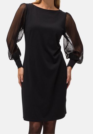 Frau trägt knielanges schwarzes Kleid mit durchsichtigen, plissierten langen Ärmeln und engen Manschetten, steht vor einfarbigem Hintergrund.