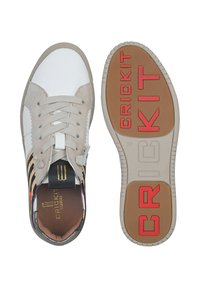 Sneaker mit einem weißen und beigen Obermaterial, schwarzen und goldenen Streifen, grauen Akzenten und einer Gummisohle mit roter "CRICKIT"-Schrift.