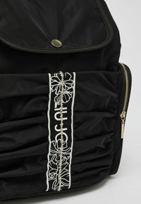 Czarny plecak z nylonu o marszczonej fakturze, z srebrnym kwiatowym detalem i złotymi akcentami. Zawiera magnesową klapkę oraz kieszeń na zamek.