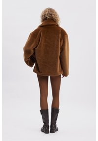 Brun fleecejacka med stor krage, boxig passform, sänka axlar och textilmaterial, i kombination med åtsittande bruna tights och svarta knähöga stövlar.