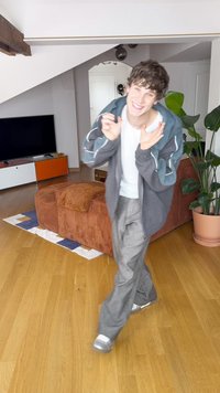 Sweater cinzento oversize com detalhes em verde-azulado, camisa branca e calças cinzentas. Ambiente interior com um sofá de bombazine castanho e plantas.