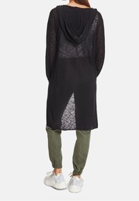 Černý dlouhý cardigan s kapucí, texturovaná pletenina, otevřený zadní díl a boční rozparky, nošený přes olivově zelené zúžené kalhoty a bílé tenisky.