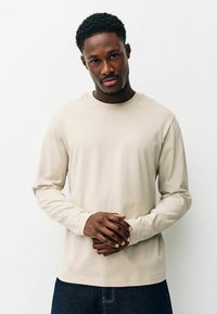 Beige pitkähihaisen t-paidan, joka on valmistettu pehmeästä kankaasta. Paidassa on pyöreä pääntie ja rento istuvuus, ja se on puettu tummien farkkujen päälle.