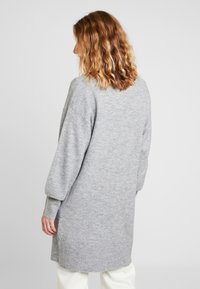 Lång grå tröja gjord av mjukt stickat material, med vida ärmar och ribbat nederkant. Designen är lös och når halvvägs ner på låren.