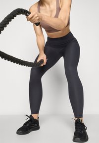 Leggings noires côtelées et un soutien-gorge de sport marron avec des bretelles larges. L'individu tient une corde de bataille noire. Chaussures de sport noires avec des lacets.