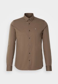 Chemise marron à manches longues boutonnée avec col et petit logo blanc "ck" sur la poitrine gauche sur fond uni.