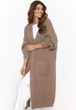 Kobieta uśmiechająca się, ubrana w długi, gruby brązowy kardigan z dzianiny na białej bluzce i białych spodniach na białym tle.