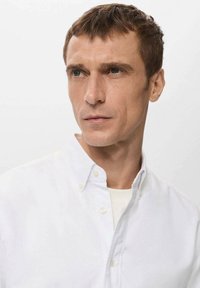 Camisa blanca abotonada con un cuello suave, que presenta una textura lisa y un diseño limpio, llevada sobre una camiseta de color claro.