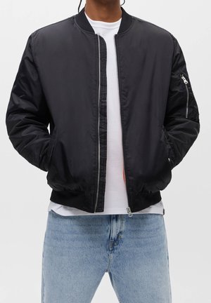 Personne portant une veste bomber noire sur un t-shirt blanc et un jean bleu clair, debout avec les mains dans les poches de la veste sur un fond uni.