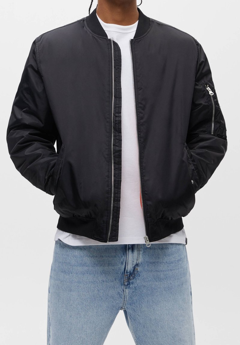 Personne portant une veste bomber noire sur un t-shirt blanc et un jean bleu clair, debout avec les mains dans les poches de la veste sur un fond uni.