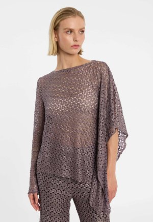 Blonde vrouw die een loszittende, mauve gehaakte top draagt met asymmetrische mouwen en bijpassende gehaakte broek, kijkt opzij.