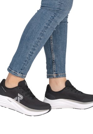 Pieds portant des baskets de sport noires avec des semelles blanches et un jean bleu ajusté, montrés en plein pas sur fond blanc.