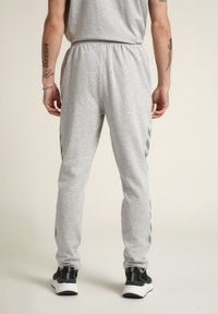 Graue Baumwoll-Jogginghosen mit elastischem Bund, schmal zulaufenden Beinen und schwarzen Seitstreifen. Getragen mit schwarzen Sportschuhen.