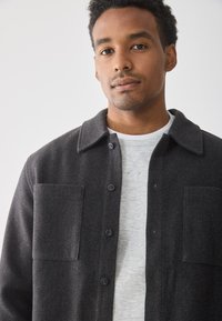 Chaqueta de camisa de lana gris oscuro con cuello, cierre de botones y dos bolsillos en el pecho, llevada sobre un sudadera gris claro.