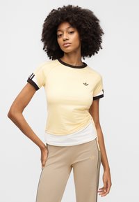 Κίτρινο αθλητικό πουκάμισο με κοντά μανίκια, καφέ ριγέ γιακά, λευκό τελείωμα και μαύρες ρίγες στα μανίκια. Διαθέτει μικρό μαύρο λογότυπο στο στήθος.