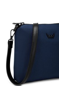 Námořnická modrá látková taška s černým koženým popruhem a akcenty. Disponuje zipovým uzávěrem a nenápadným logem na přední straně. Kompaktní obdélníkový tvar.