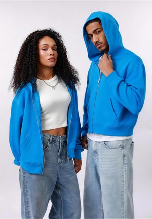 Fiatal nő göndör hajjal, kék cipzáras pulóverben és rövid fehér topban áll egy kék kapucnis pulóvert és világos farmert viselő férfi mellett, fehér háttér előtt.