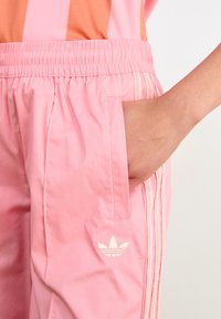 Pantalones deportivos rosas con cintura elástica, bolsillos laterales y tres rayas color crema. Cuenta con un logo bordado en el bolsillo izquierdo.