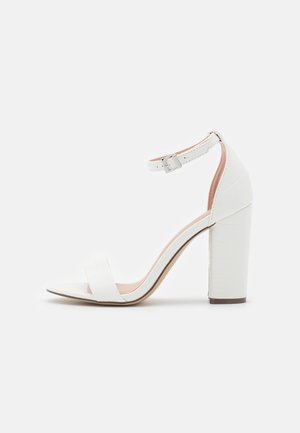 Hvid højhælet sandal med ankelrem, blokhæl og åben tå på en ensartet lys baggrund.
