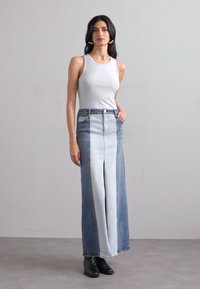 Denim maxi skirt with light and dark blue panels, featuring a front slit and a high waist. Worn with a fitted white tank top and black boots.
