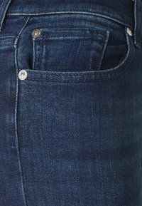Jeans en denim d'un indigo foncé, avec une poche avant accentuée par des boutons argentés et une légère décoloration sur le tissu pour une texture.