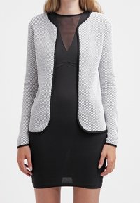 Czarna sukienka o dopasowanym kroju, uzupełniona białym, strukturalnym kardiganem z heksagonalnym wzorem i czarnym obszyciem.