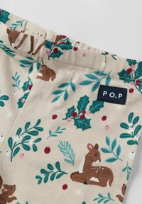 Close-up van beige stof met groene hulstbladeren, rode bessen en bruine hertenpatronen, met een klein "P.O.P" label en een elastische tailleband.