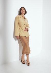 Beige long-sleeve shirt with buttons, paired with a sheer brown skirt and black flip-flops, leaning against a white wall.