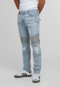 Világoskék farmernadrág, amelynek kopott területei vannak, és csíkozott szövetbetétek díszítik a térdeknél. Slim fit fazonú, szabványos öt zsebes kialakítással.