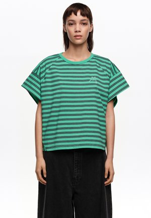 Joven con una camiseta holgada verde con rayas horizontales negras y pantalones negros de pierna ancha, de pie frente a un fondo blanco.