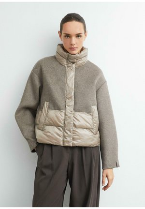 Junge Frau trägt eine beige und taupe gepolsterte Winterjacke mit hohem Kragen und dunkle, locker sitzende Hose und steht vor weißem Hintergrund.