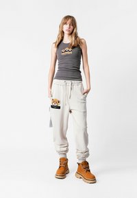 Camiseta sin mangas gris con osos bordados, pantalones de chándal beige con parche de oso y bolsillo, y botas marrones con cordones y suelas gruesas.