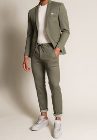 Olive green casual suit with a tailored blazer, elastic waistband trousers, and white sneakers. Textured fabric; pocket square detail.