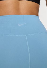 Világoskék, magasan szabott leggings sima felülettel, lapos varrásokkal és egy kis visszaverődő Nike logóval a derekán.