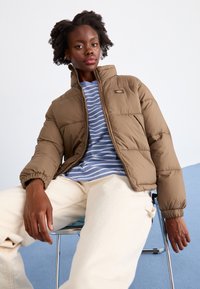 Giacca a vento marrone con design trapuntato e colletto alto, indossata sopra una camicia a righe blu. Modello seduto su una sedia con pantaloni color crema.