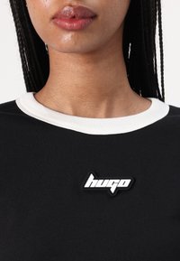 Svart plagg med en hvit ribbestrikket krage. Har et gummiaktig logo-patch som sier "hugo" i hvitt på brystet. Glatt tekstur, uformell design.