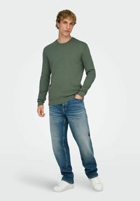 Chemise texturée verte à manches longues avec encolure ronde portée avec un jean bleu et des baskets blanches. Le mannequin est debout sur un fond neutre.