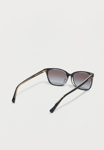 Μαύρα και χρυσά ορθογώνια γυαλιά ηλίου με φακούς σε σκούρο διαβαθμισμένο γκρι και το λογότυπο "RALPH" στα πλαινά, ακουμπισμένα σε λευκή επιφάνεια.