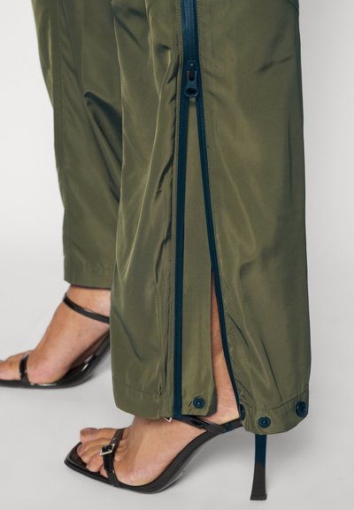 Olijfgroene broek met een gladde textuur, voorzien van een zijrits en knoopdetails, gecombineerd met zwarte sandalen met hoge hakken en bandjes.