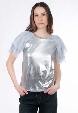 Zilverkleurige metallic top met doorschijnende grijze tule ruches aan de mouwen, een ronde halslijn en een gladde textuur. Draagt donkere spijkerbroeken.