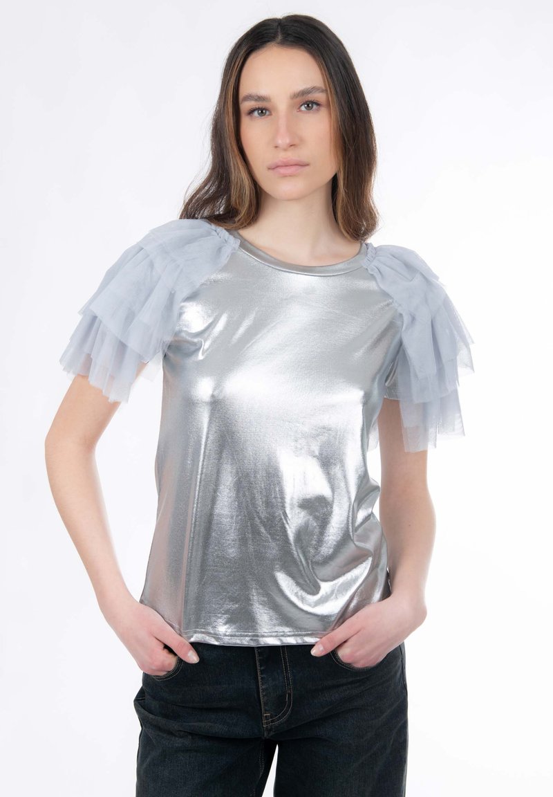 Top metálico plateado con mangas voluminosas de tul gris translúcido, escote redondo y textura suave. Combinado con vaqueros oscuros.