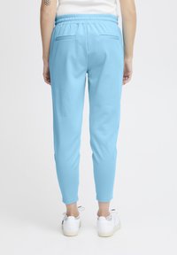 Persoon in lichtblauwe elastische broek en witte sneakers, van achteren gezien tegen een plain witte achtergrond.