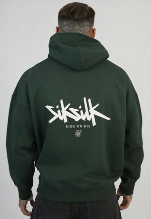 Mężczyzna z krótkimi, wzorzystymi włosami nosi ciemnozieloną bluzę z kapturem z napisem „SikSilk Ride or Die” na plecach, stojąc na jednolitym tle.