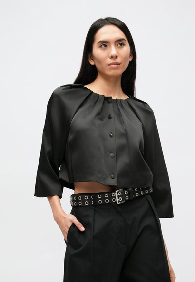 Schwarze, gekürzte Bluse mit rundem Ausschnitt, plissiertem Design und Knopfverschluss. Kombiniert mit hoch taillierter Hose und einem Nietengürtel.